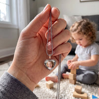 Thumbnail for The silver heart photo pendant held up, real small size, her little girl playing behind | SKU: TPH-CHTN-SILVER-SMALL
