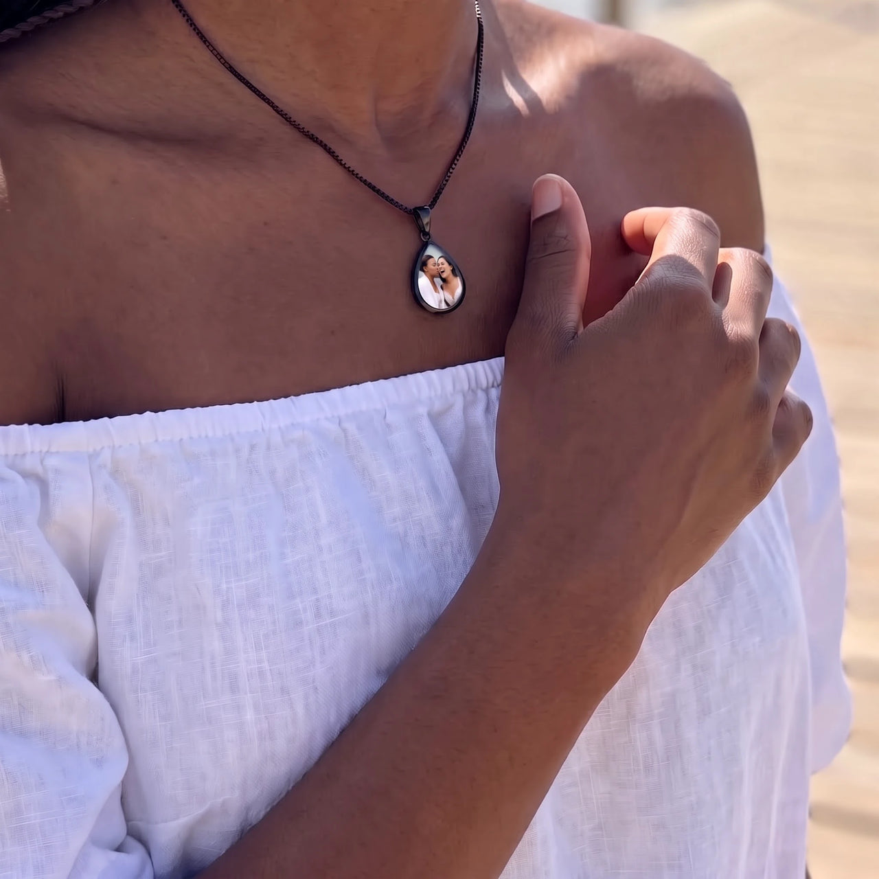 Woman in an off-shoulder white top touching the black teardrop pendant to reveal the photo | SKU: TPH-DRPN-BLACK