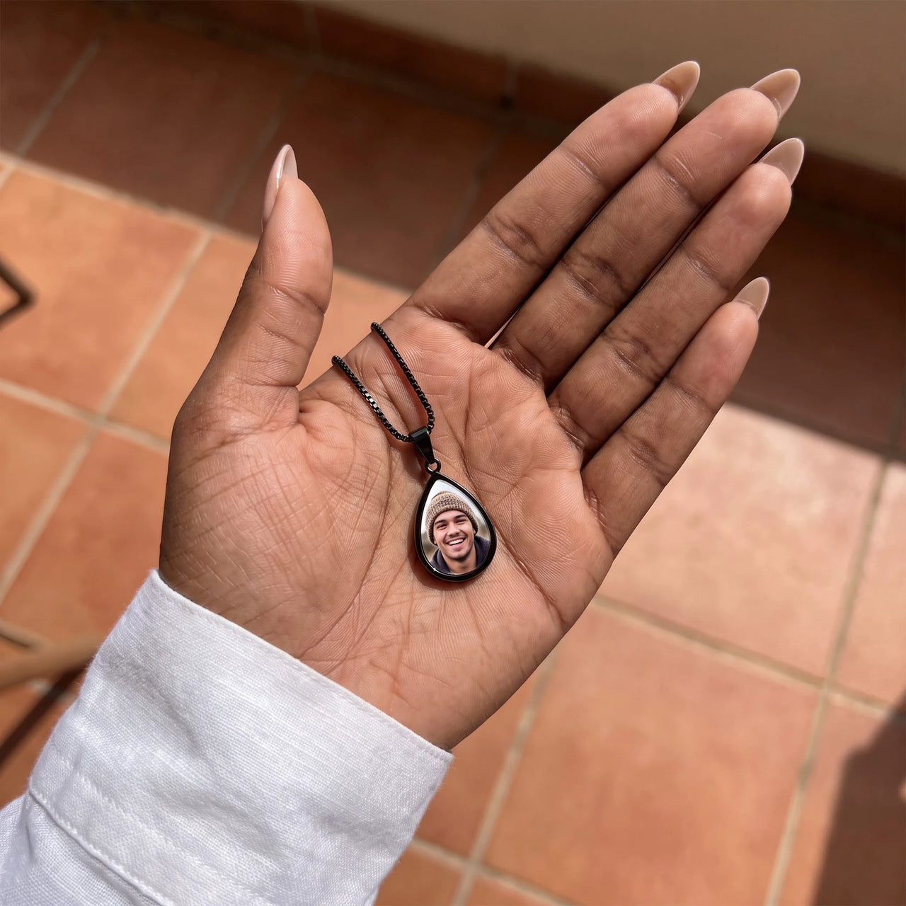 The black teardrop photo pendant in an open palm over terracotta tiles, showing its real small size | SKU: TPH-DRPN-BLACK