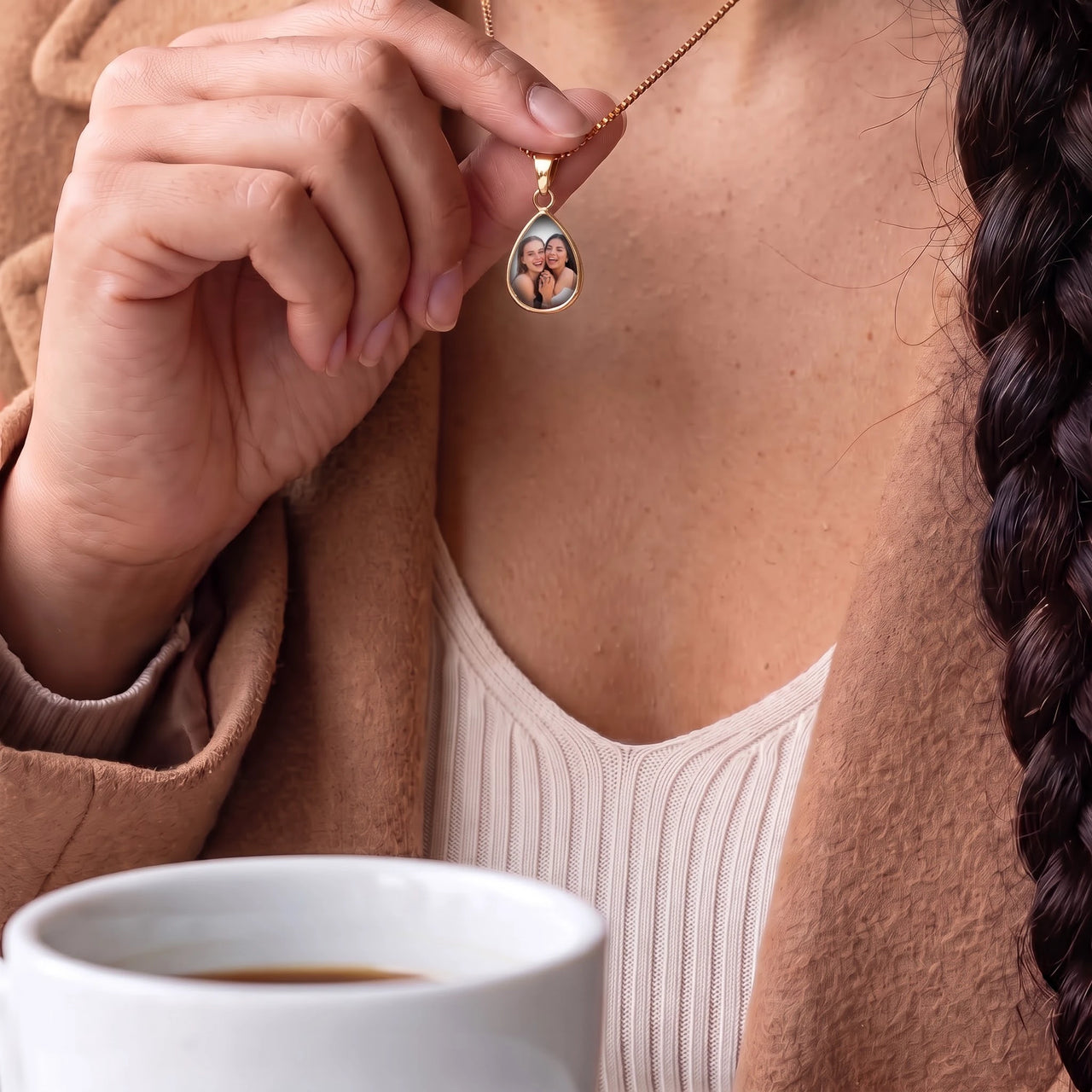 Woman touching the gold teardrop pendant to reveal the photo, a cup of coffee in front of her | SKU: TPH-DRPN-GOLD