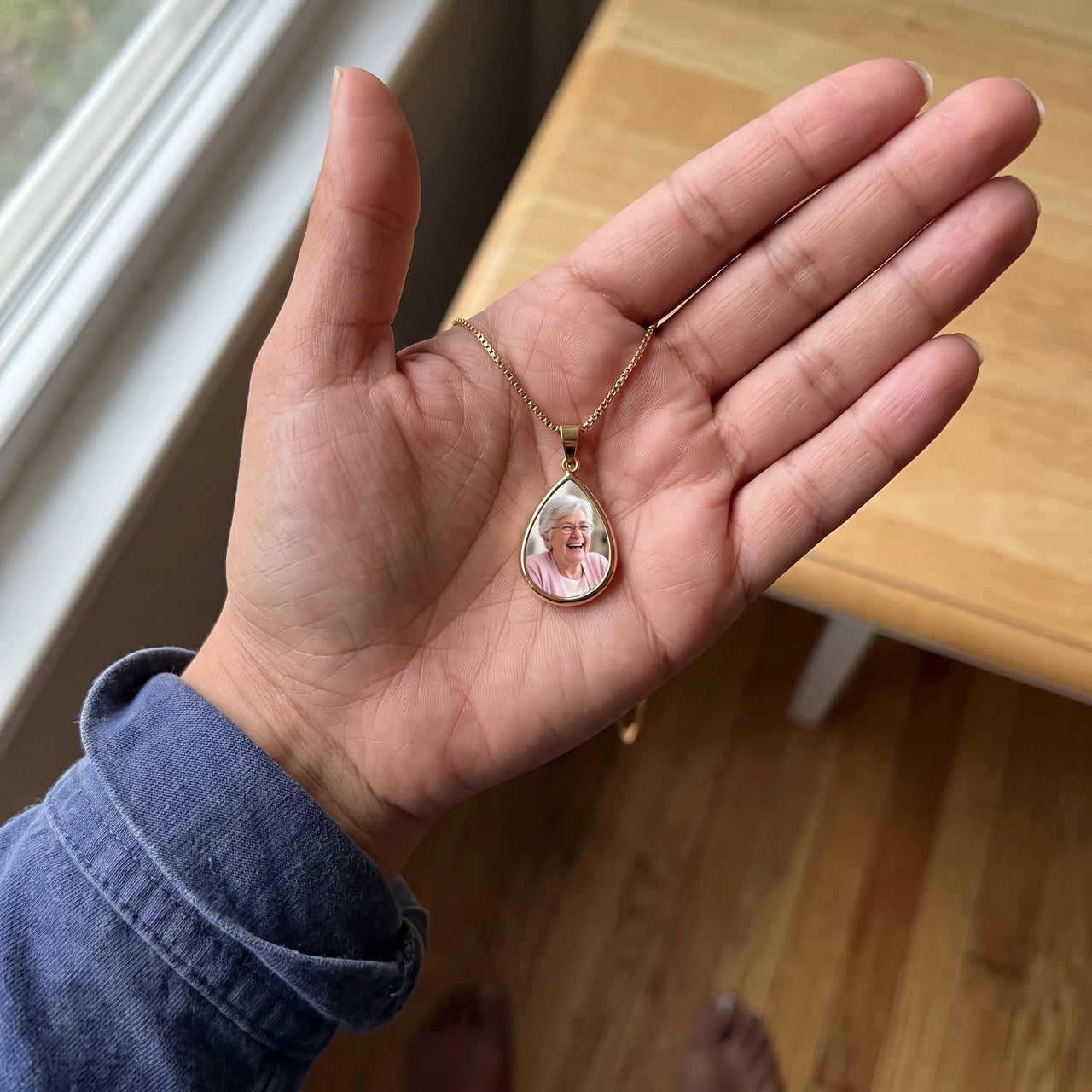 The gold teardrop photo pendant in an open palm by a window, showing its real small size | SKU: TPH-DRPN-GOLD