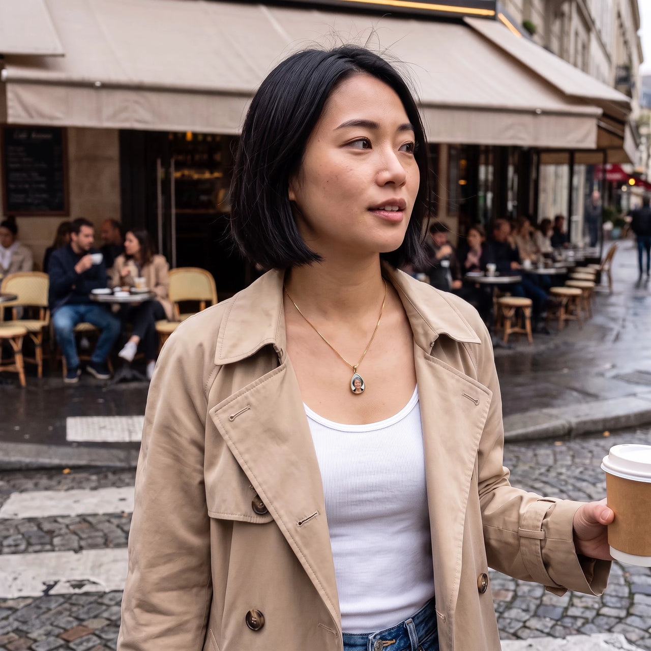 Woman in a trench coat with a coffee by a Paris café, wearing the gold secret drop photo necklace | SKU: TPH-DRPN-GOLD